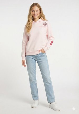 Lächelnde Frau trägt hellrosa Hoodie mit Aufnähern, hellblaue Jeans und weiße Turnschuhe, steht mit einer Hand in der Tasche.