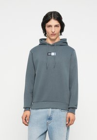 Felpa con cappuccio grigia in tessuto morbido, dotata di tasca a marsupio e patch con logo frontale. Maniche lunghe con polsini e orlo a costina.