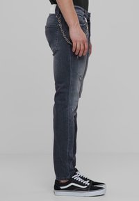 Tummat farkut, joissa on kulutuspinnat, slim fit -malli ja näkyvä ketjuakseli. Yhdistetty mustiin lenkkareihin, joissa on valkoiset raidat.