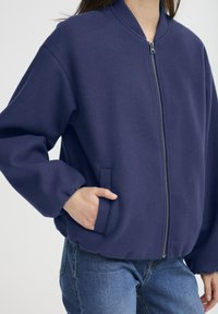 Jūra bomber jakas izgatavota no teksturēta auduma, ar zīmuļa priekšu, ribotām malām un sānu kabatām, kombinēta ar džinsu biksēm.