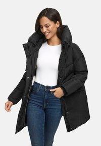 Black, puffy winter parka with a high collar, snap buttons, side pockets, and a zipper. Worn over a white top and dark jeans.