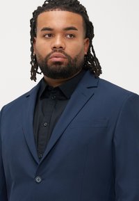 Donkerblauw colbert met een enkele knoop, inkeping revers en een borstzak. Gedragen over een zwart overhemd met knoopsluiting. Fijne textuur zichtbaar.