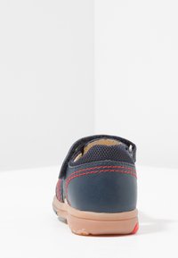 Zapato de cuero azul marino con parte superior de malla, acentos de costura roja y suela de goma flexible; cuenta con un cierre de gancho y lazo para facilitar el uso.
