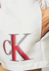 Pantaloni corti bianchi con un grande logo "ck" rosso e nero a pois sul lato. Texture liscia, vestibilità rilassata e tasche laterali.