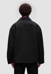 Sort oversized jakke med klassisk krave og sænkede skuldre. Har en glat tekstur og minimalistisk design, ingen synlige lukninger.