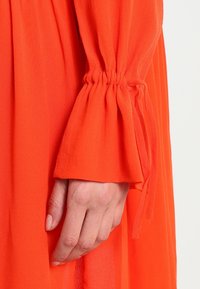Pitkähihainen, kirkkaan oranssi mekko, jonka hihansuissa on rypytetty kangas. Mekko on pehmeää, kevyttä materiaalia ja virtaavaa muotoilua.