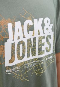 Žalias medvilninis marškinėliai su dideliu baltu tekstu "JACK & JONES" ir geltonais bei oranžiniais miesto žemėlapių grafika apačioje, trumpomis rankovėmis, laisva forma.