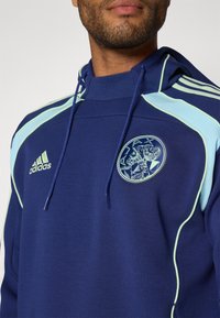 Mørkeblå hettegenser laget av myk stoff, med lyseblå kanting, en sentral logo-patch, og justerbare snorer i hetta.
