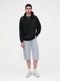 Jeune homme debout portant un sweat à capuche noir avec logo, un short bleu clair arrivant aux genoux, des chaussettes blanches et des baskets blanches sur fond uni.