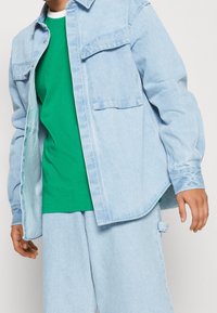 Light blue denim shirt with two front pockets, layered over a green t-shirt, paired with light blue pants, casual and relaxed fit.