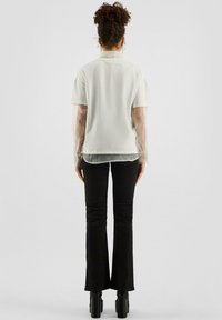 Witte top met korte mouwen en doorschijnende lange mouwen, gecombineerd met zwarte flared broek en chunky haklaarzen, gezien van achteren.