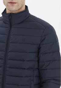 Marinblå puffjacka med hög krage, quiltat mönster, framdragkedja och liten logotyp på bröstet.