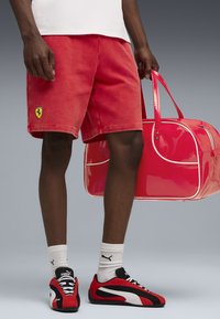 Mand iført røde Ferrari-shorts og røde Puma-sneakers, holder en blank rød duffelbag med hvid piping foran en grå baggrund.
