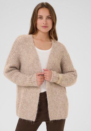 Une femme porte un cardigan en maille beige à la texture travaillée sur un haut blanc et un pantalon foncé, et le tient ouvert sur le devant devant un fond clair et uni.