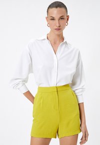 Camisa blanca de cuello con mangas largas, combinada con pantalones cortos amarillos brillantes de talle alto, texturizados, que cuentan con bolsillos laterales y un dobladillo limpio.