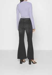 Kvinde iført sorte udsl åede jeans, sorte højhælede sandaler og en langærmet lavendelfarvet top, stående med front mod en hvid væg.