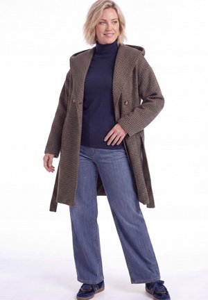 Blond woman wearing a brown checkered coat, navy turtleneck, wide-leg blue jeans, and blue sneakers standing against white background.