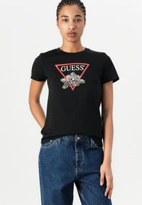 Svart T-skjorte i bomull med en rød trekantet logo som har ordet "GUESS" og et blomsterdesign. Kort erm og rund hals.