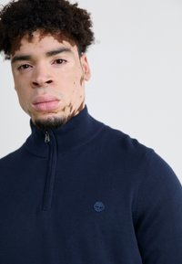 Mornarica zip-vest sweater s pletenom teksturom, rebrastim ovratnikom i malim plavim logotipom drveta na prsima.