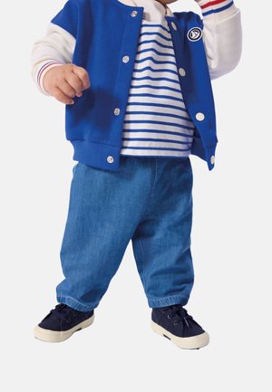 Småbarn iført blå og hvid varsityjakke, stribet skjorte, løstsiddende blå jeans og marineblå sko står med den ene arm hævet.