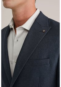 Blazer blu navy con una trama sottile e revers a punta, abbinato a una camicia color grigio chiaro con colletto e un bottone decorativo sul blazer.