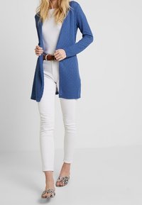 Cardigan de punto azul, con frente abierto, mangas largas y un patrón texturizado, combinado con jeans blancos y sandalias con estampado.