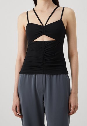 Femme portant un haut noir froncé sans manches à fines bretelles avec une découpe sous la poitrine, associé à un pantalon gris ample.
