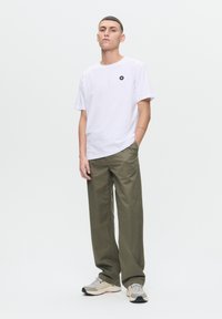 Weißes Baumwoll-T-Shirt mit kleinem schwarzem Logo, kombiniert mit lockeren olivgrünen Hosen und beigen Sneakers mit grauen und schwarzen Akzenten.