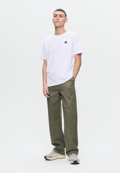 T-shirt en coton blanc avec un petit logo noir, associé à un pantalon large vert olive et des baskets beiges avec des accents gris et noirs.