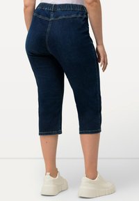 Tummansinisen väriset denimpituiset capri housut, joissa on joustava vyötärö, lyhennetty pituus ja näkyvät ompelusaumat helmassa. Yhdistettynä valkoisiin tennareihin.