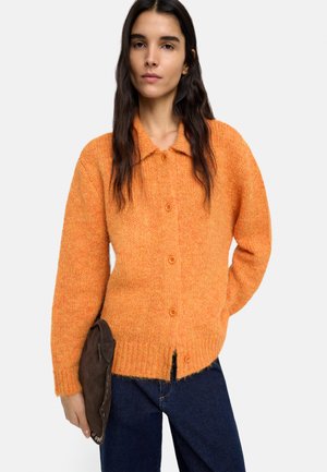 Kobieta ubrana w pomarańczowy rozpinany sweter z kołnierzem, ciemne dżinsy, trzyma brązową zamszową kopertówkę na jednolitym tle.