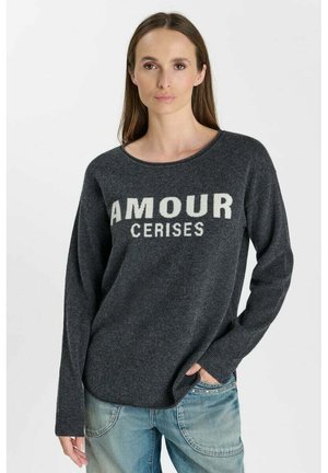 Vrouw met lang bruin haar die een donkergrijze trui draagt met de tekst "AMOUR CERISES" en lichtblauwe spijkerbroek, staand met één hand in de zak.