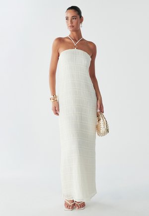 Woman in a white strapless maxi dress with a textured pattern, holding a gold woven clutch, posing against a light background.