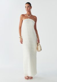 Witte strapless maxijurk met gestructureerde stof, voorzien van een rechte halslijn en subtiele verticale lijnen. Gecombineerd met een gevlochten gouden handtas.