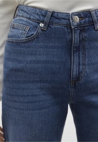 Tmavě modré džíny z denimu s hladkým povrchem, které mají přední kapsu, kovové zapínání na knoflík a prošívané detaily podél švů.