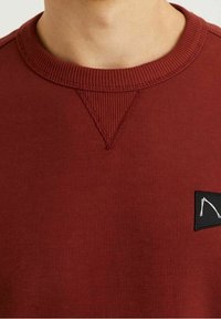 Sweat-shirt bordeaux avec un col côtelé, un détail de surpiqûre en V et une petite étiquette de logo noire sur le côté gauche. Tissu doux et texturé.