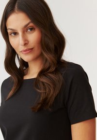 Haut noir à manches courtes avec un décolleté carré, texture lisse et silhouette ajustée, porté par un mannequin avec des cheveux bruns ondulés.
