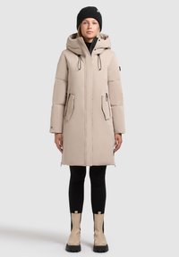 Manteau doudoune beige avec capuche, fermeture éclair à l'avant et boutons-pression, deux poches latérales et accents noirs. Porté avec des leggings noirs et des bottes beige à la cheville.