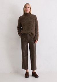 Bruin, geweven trui met hoge hals, gecombineerd met bruine leren recht-pas broek en donkerbruine instappers. Effen achtergrond.
