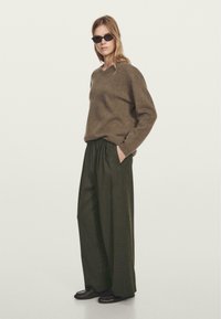 Maglione di lana marrone con scollo a V, abbinato a pantaloni larghi verde scuro. Indossato con occhiali da sole neri rettangolari e scarpe marroni.