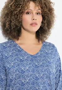Donna con capelli ricci che indossa orecchini a cerchio argentati e una maglietta blu con un motivo floreale a V, su uno sfondo bianco.