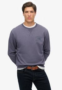 Lilla sweatshirt med rund halsudskæring og ribstrikkede manchetter. Den har et lille "Superdry Athletics" logo i mørkeblå på venstre brystområde.