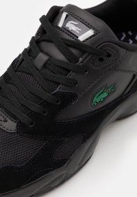 Schwarzer Sneaker aus einer Kombination von Leder und Mesh-Materialien, mit einem auffälligen grünen Krokodil-Logo und strukturierten Akzenten.