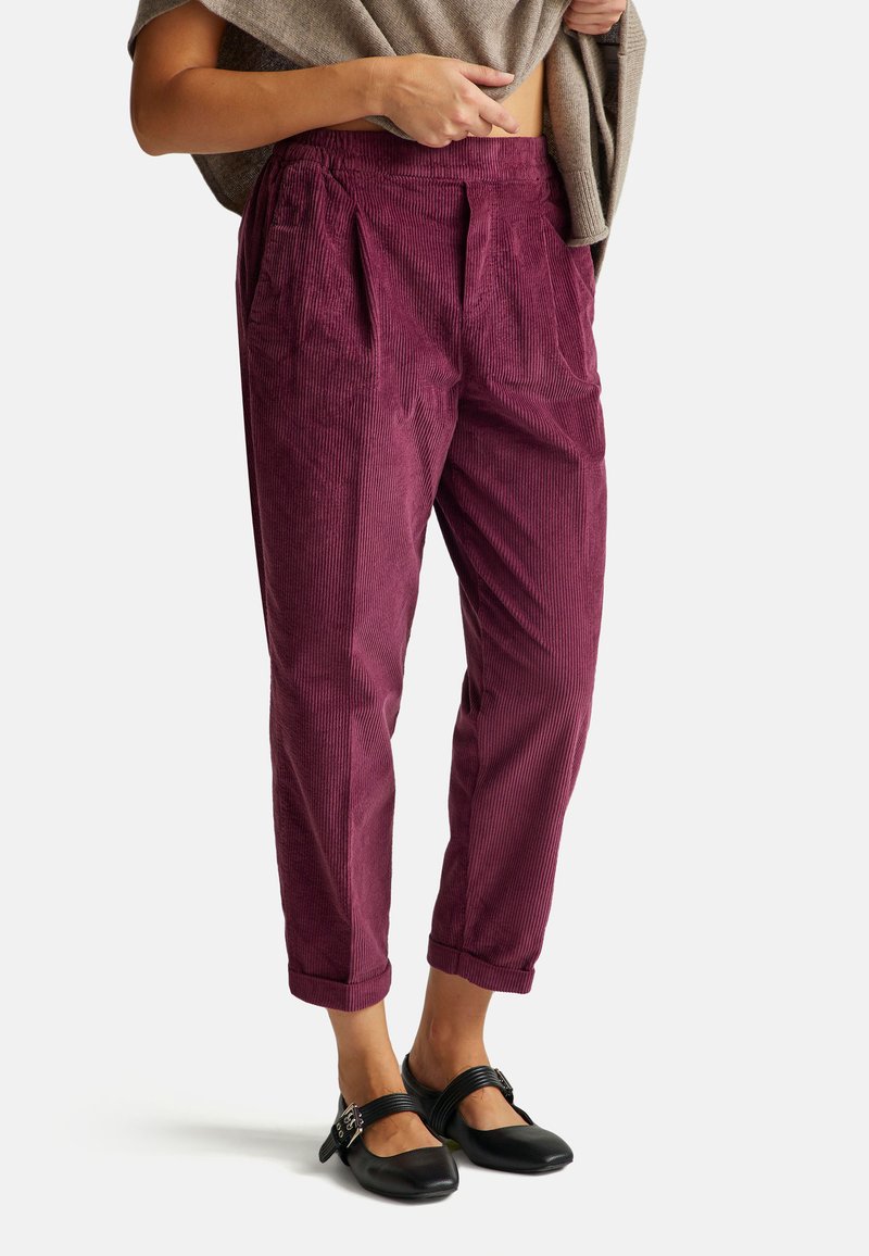 Person wearing high-waisted purple corduroy pants and black Mary Jane shoes, holding a beige sweater around the shoulders.