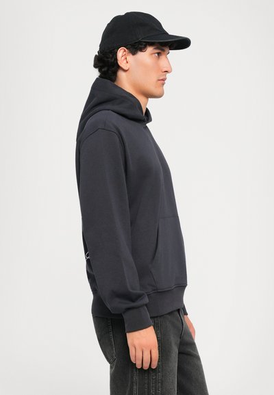 Sudadera gris oscuro con bolsillo canguro, corte holgado y dobladillo acanalado. Combinada con gorra negra y jeans de mezclilla oscuros. Vista lateral.