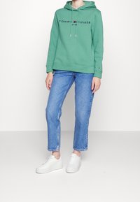 Osoba v zelené mikině Tommy Hilfiger, modrých džínách a bílých teniskách, stojící na pozadí jednoduchého světlého pozadí.