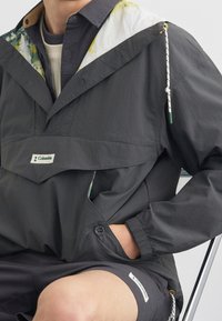 Personne portant une veste Columbia gris foncé avec une doublure de capuche blanche et jaune et un short assorti, assise avec une main dans la poche de la veste.