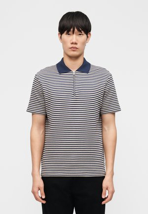 Jeune homme portant un polo à manches courtes rayé bleu marine et blanc avec un col bleu marine et une fermeture éclair à l'avant, se tenant devant un fond uni.