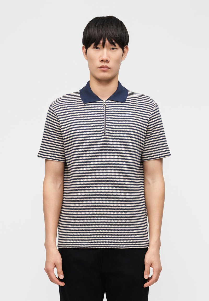 Νέος άνδρας που φοράει μπλε σκούρο και λευκό ριγέ κοντομάνικο πόλο με μπλε σκούρο γιακά και φερμουάρ στο μπροστινό μέρος, στέκεται μπροστά σε ένα απλό φόντο.