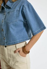 Helles blaues, verkürzt geschnittenes Jeanshemd mit kurzen Ärmeln, einer Brusttasche und silbernen Knöpfen; kombiniert mit beigen Hosen und einem braunen Armreif.
